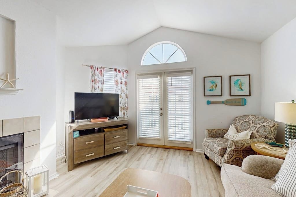 A living room with a television, a fireplace, a patterned sofa, and a large window with blinds.