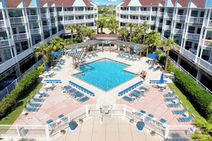 Ein Hotel mit einem zentral gelegenen Schwimmbad, umgeben von Liegestühlen und Sonnenschirmen, mehreren Balkonen und Palmen.