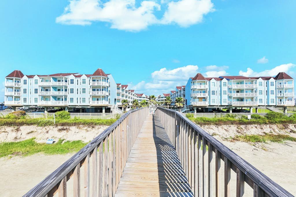 A wooden boardwalk leading to beachfront condos with balconies, under a clear sky.