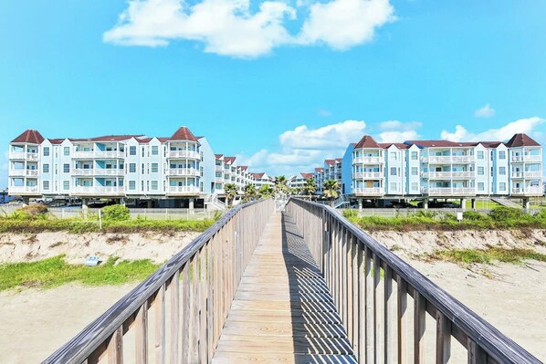 Une promenade en bois mène à des appartements situés en bord de mer, dotés de balcons, sous un ciel dégagé.