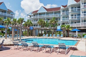 A hotel with a swimming pool, lounge chairs, and palm trees. - Seascape Condos #1201 (Galveston)