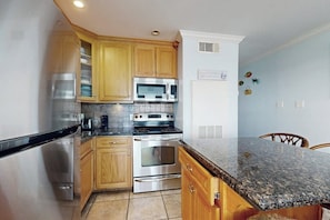 A kitchen with wooden cabinets, stainless steel appliances, a granite countertop, and a white wall with decorative items. - Seascape Condos #1201 (Galveston)