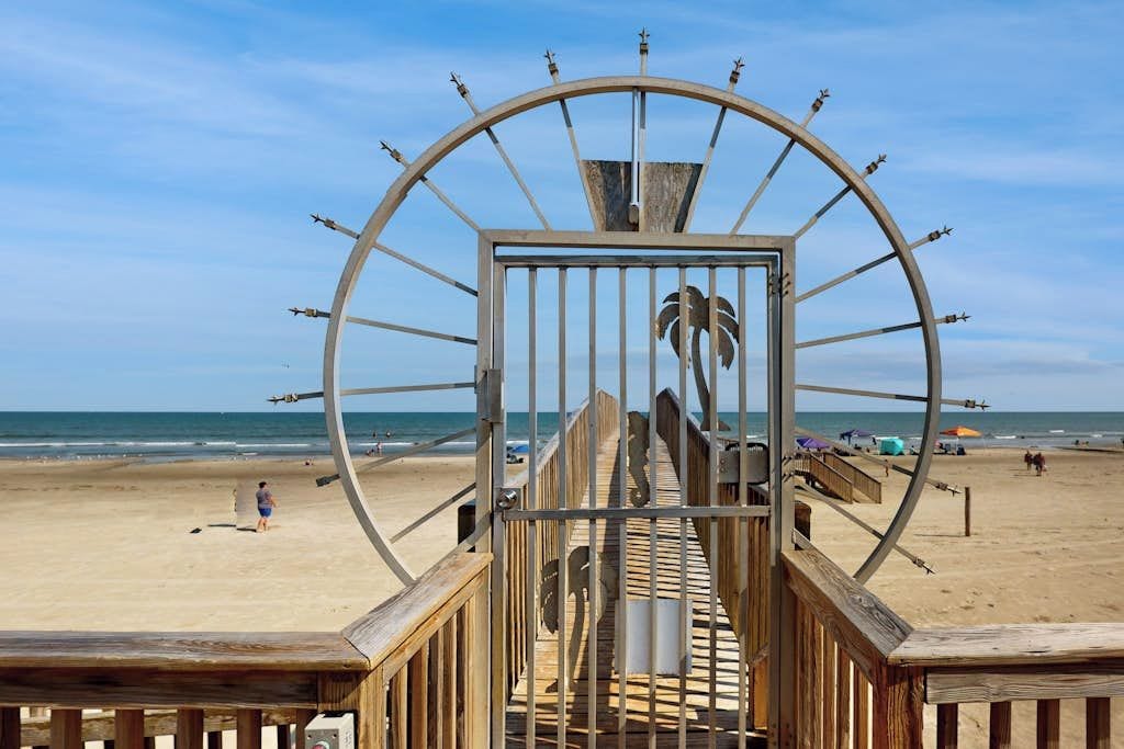 A beach with a wooden walkway leading to a large metal gate with a palm tree design.