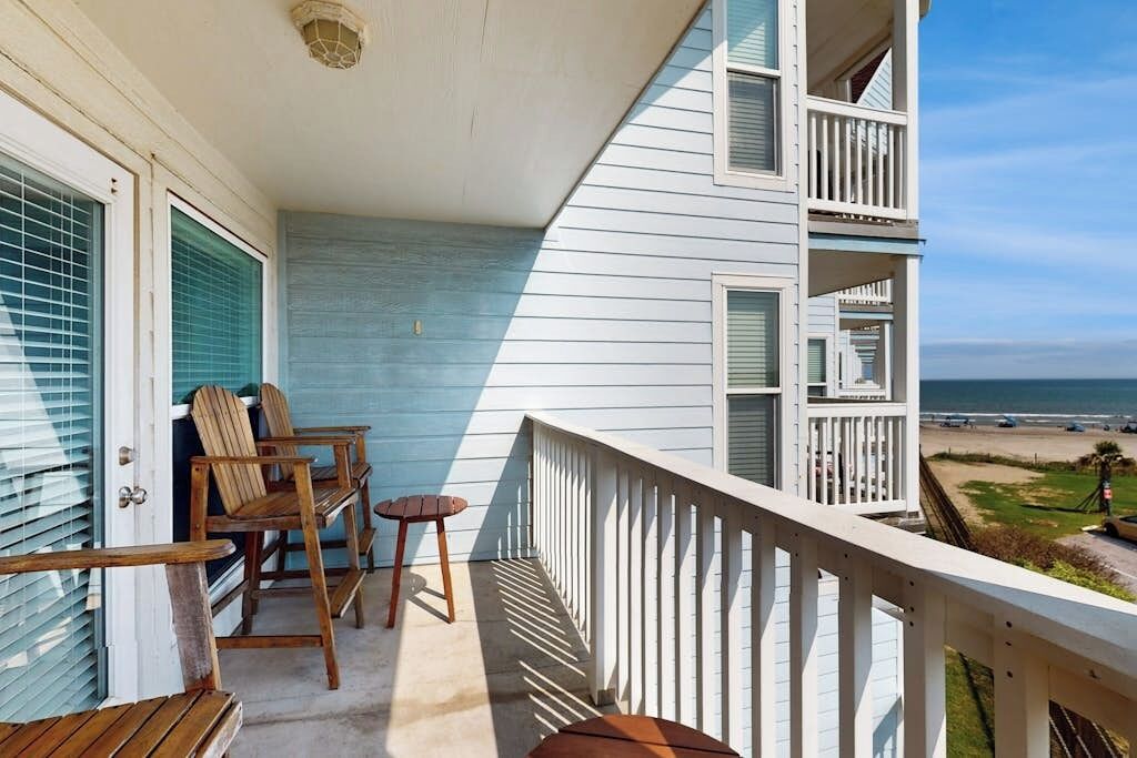 A balcony with a view of the beach, wooden furniture, and a white railing.