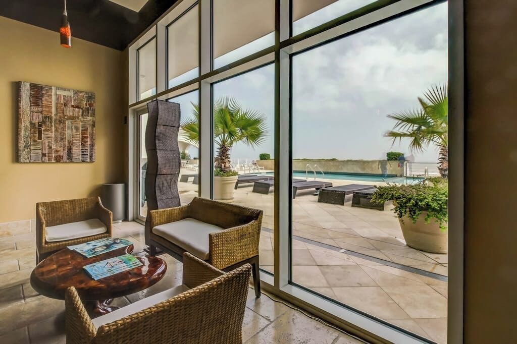 Quarto com janelas amplas, móveis de vime, uma pequena mesa e vista para área de piscina externa.