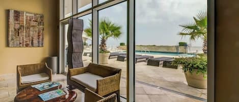 A room with large windows, wicker furniture, a small table, and a view of an outdoor pool area.