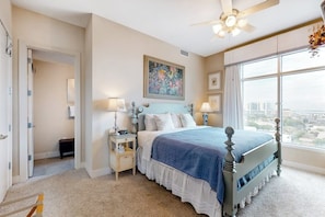 A bedroom with a bed, bedside tables, a ceiling fan, and a large window with a city view. - Emerald by the Sea #806 (Galveston)