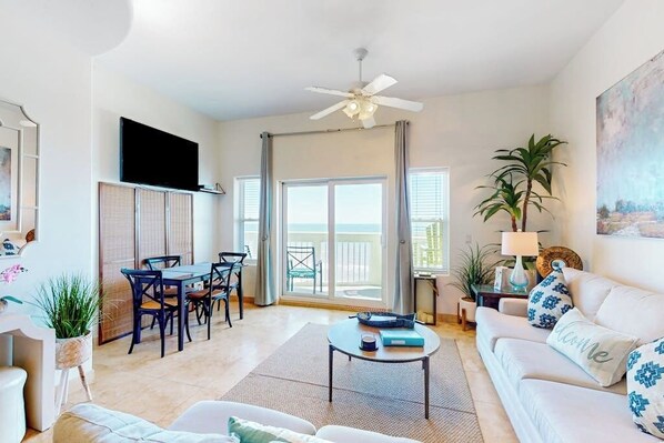 A living room with a sofa, coffee table, TV, dining area, and a balcony with a view. - Seaside Oasis - Gulf Views, Pool & Fishing Pier (Galveston)