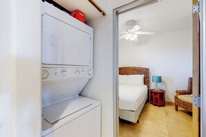 A hotel room with a washing machine, a bed with white bedding, a nightstand with a lamp, and a chair. - Seaside Oasis - Gulf Views, Pool & Fishing Pier (Galveston)