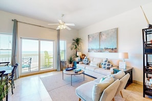 A living room with a sofa, coffee table, and a view of the outdoors through a sliding glass door. - Seaside Oasis - Gulf Views, Pool & Fishing Pier (Galveston)