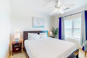 A hotel room with a bed, a nightstand, a ceiling fan, a window with curtains, and a painting on the wall. - Seaside Oasis - Gulf Views, Pool & Fishing Pier (Galveston)