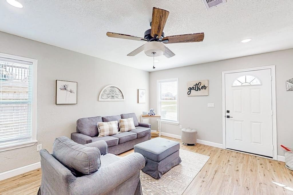 Ein Wohnzimmer mit grauem Sofa, Hocker, Deckenventilator und einer Tür mit dem Schild 'gather'.
