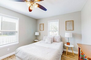 A bedroom with a bed, two bedside tables, a ceiling fan, and two windows with blinds. - Out of the Blue - Fire Pit & Patio Near the Beach (Galveston)