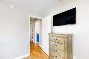 A hallway with a wooden floor, a white wall, and a television mounted above a wooden dresser. - Out of the Blue - Fire Pit & Patio Near the Beach (Galveston)