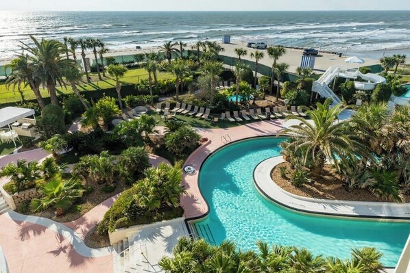 Zwembad van een resort met een golvend ontwerp, omgeven door palmbomen en ligbedden, met uitzicht op de oceaan.