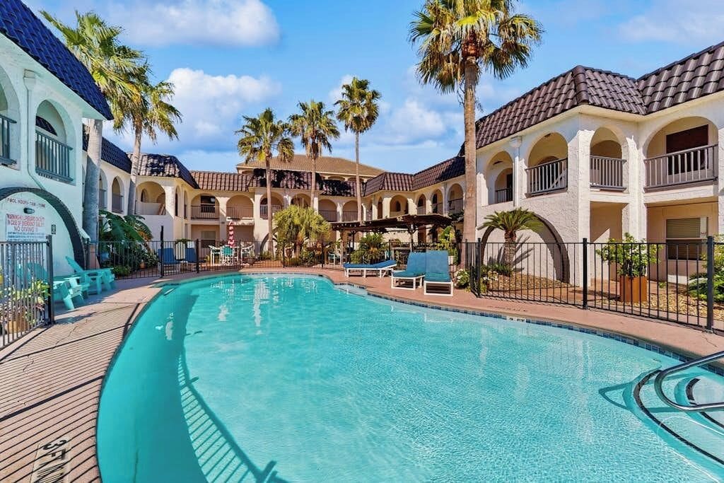A pool area with a clear blue pool, lounge chairs, and a building with arched balconies and palm trees.