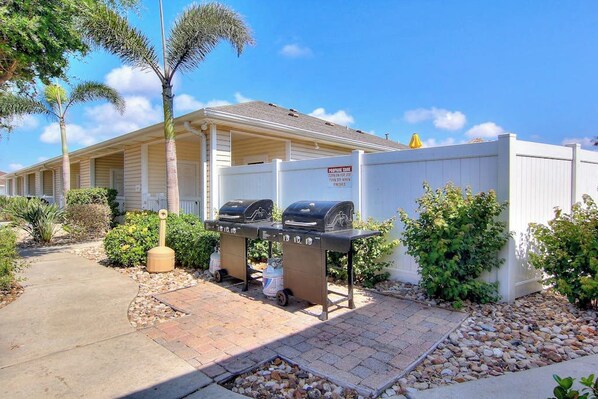 A motel with a barbecue area, a covered walkway, and a fenced yard with palm trees. - Palm Bay 202 - Heated Pool, BBQ & Beach Access (Corpus Christi)