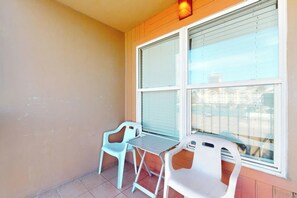 Ein Balkon mit zwei Stühlen und einem Tisch, ein Fenster mit Jalousien sowie eine Wand mit einer Lampe.