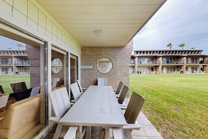 Et udendørs spiseområde med et langt bord og stole, en murstensvæg med et rundt vindue samt udsigt til en bygning med balkoner.