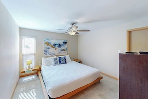 A bedroom with a bed, a ceiling fan, a painting, a nightstand, and a desk. - Sunchase Beachfront #130 (South Padre Island)