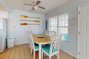 Un comedor con una mesa de madera y sillas blancas, un ventilador de techo y una ventana con persianas.