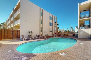 Una piscina con sillones de descanso y un patio de ladrillo, rodeada por un edificio con balcones.