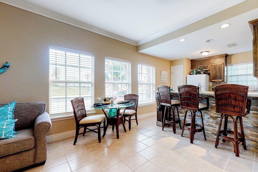 A spacious dining area with a round table and multiple chairs, a kitchen with wooden cabinets, and large windows providing natural light.