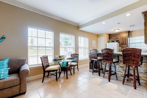 A spacious dining area with a round table and multiple chairs, a kitchen with wooden cabinets, and large windows providing natural light. - Sun Dancer Condominiums 1 (South Padre Island)