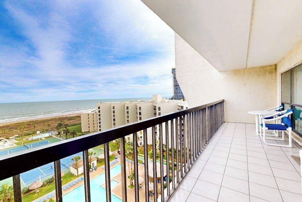 A balcony with a view of the beach and ocean, a pool, and a dining area with chairs.