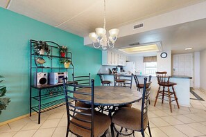 A dining area with a round table, chairs, a chandelier, and a kitchen in the background. - Saida Towers III #3116 (South Padre Island)