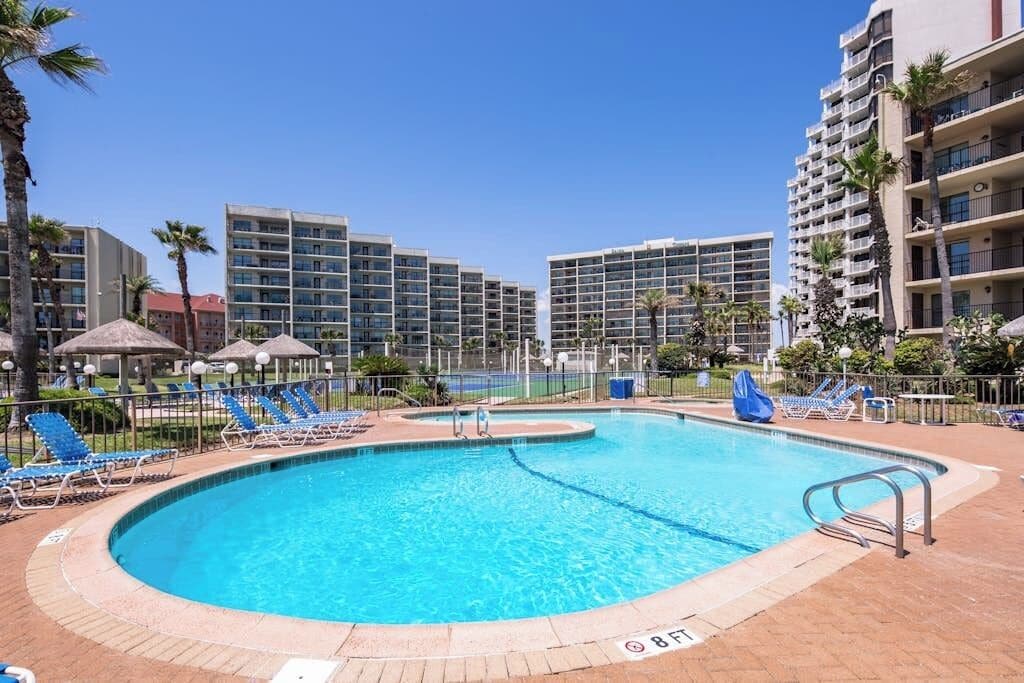 A swimming pool with blue water, surrounded by lounge chairs and umbrellas, with high-rise buildings in the background.