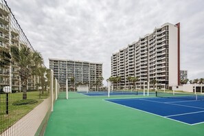 A tennis court with a net, surrounded by a fence and palm trees. - Saida Towers III #3116 (South Padre Island)