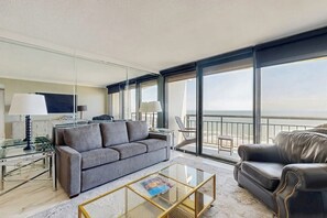 Ein modernes Hotelzimmer mit einem grauen Sofa, einem Ledersessel, einem Glastisch und einem großen Fenster mit Blick aufs Meer.
