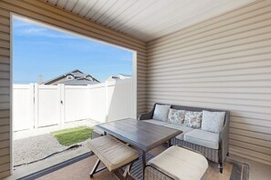 Eine überdachte Terrasse mit Tisch und zwei Stühlen, ein Korbsessel mit Kissen sowie ein Blick auf einen weißen Zaun und Häuser im Hintergrund.