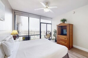 A hotel room with a bed, a wooden cabinet, a ceiling fan, and a large window. - Sunchase IV #525 - Gulf Views, Pools & Sauna (South Padre Island)