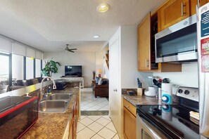 A modern kitchen with stainless steel appliances, a dining area with a TV, and a living room with a sofa. - Sunchase IV #525 - Gulf Views, Pools & Sauna (South Padre Island)