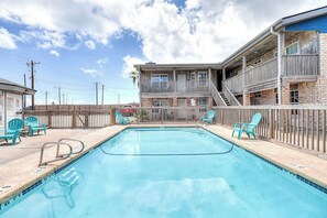 Ein rechteckiger Swimmingpool mit Holzdeck und Stühlen, umgeben von einem Zaun, mit einem Gebäude mit Balkonen und einer Treppe im Hintergrund.