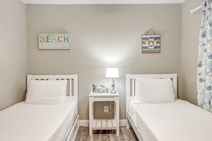 Une chambre avec deux lits simples, une table de chevet avec une lampe et des éléments de décoration murale, notamment un panneau indiquant « PLAGE » et une œuvre sur le thème de la marine.