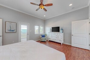 A bedroom with a bed, a ceiling fan, a dresser, a television, and a door. - Lilac Sky - Water Views, Pool & Dogs OK (Port Aransas)