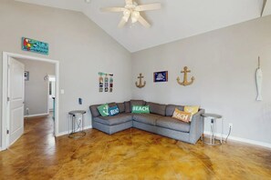 Ein Zimmer mit grauem Ecksofa, einem Deckenventilator und Wanddekorationen im maritimen Stil.