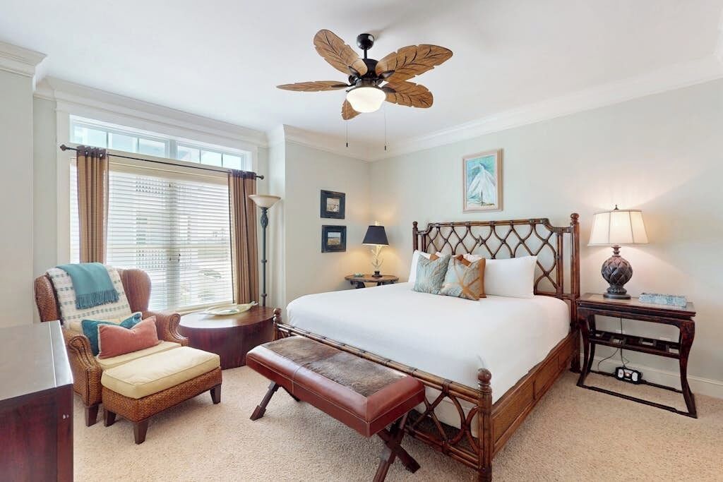 A bedroom with a bed, a chair, a bench, a nightstand, a lamp, a ceiling fan, and a window with curtains.