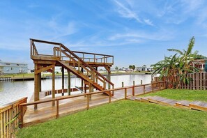 Une terrasse en bois avec un escalier menant à une plateforme surplombant une étendue d’eau, le tout entouré d’une zone herbeuse et d’une clôture en bois.