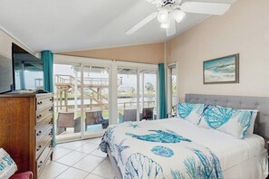 A bedroom with a bed, a ceiling fan, a sliding glass door leading to a balcony, and a painting on the wall. - The Seahorse Cottage (Corpus Christi)