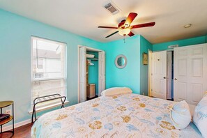 Ein Schlafzimmer mit Bett, Deckenventilator, Fenster mit Jalousien, einer Tür und einem kleinen Tisch.