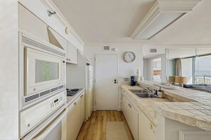 A narrow kitchen with white cabinetry, a microwave, and a stove. There is a clock on the wall and a window with a view. - Gone Gulfin' - Gulf-Front Resort, Pool & Hot Tub (Galveston)