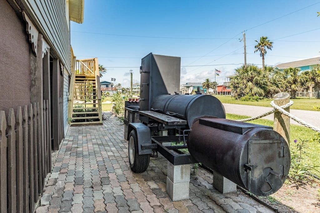 A portable barbecue grill with a chimney, placed on a paved area with a wooden fence and a building in the background.