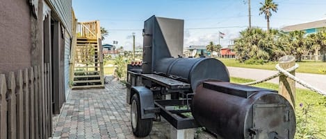 Churrasqueira portátil com chaminé, colocada em área pavimentada com cerca de madeira e um edifício ao fundo.