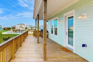 A covered wooden deck with a view of a light blue building and other structures in the background. - The Green Palm - Bay-View Patio & Shared Pool (Galveston)