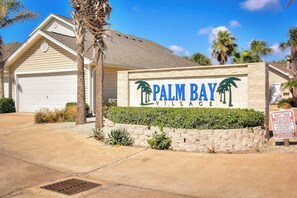 A residential property named Palm Bay Village with a garage, palm trees, and a clear sky. - Palm Bay 306 - Pool & Patio, Short Walk to Beach (Corpus Christi)