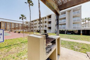 A courtyard with a barbecue grill, a fenced tennis court, and a multi-story building with balconies. - Gulf Point Condominiums #1314 (South Padre Island)